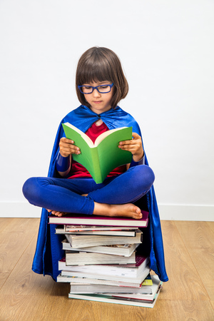 wise 6-year old schoolgirl with a super hero costume reading seated on top of a stack of books reading with eyeglasses for girl power in education and female knowledge, indoor conceptの写真素材