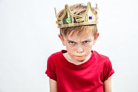 irritated beautiful spoiled boy with frowning freckles and a dirty look wearing a golden crown for mad attitude facing parenthood and education, white background, indoorsの写真素材