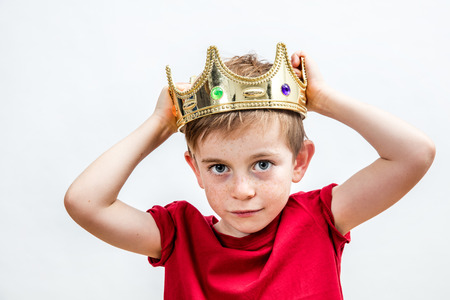 happy education and childhood concepts with an adorable 7-year old boy holding a golden king crown on his head as a wise spoiled child, white backgroundの写真素材
