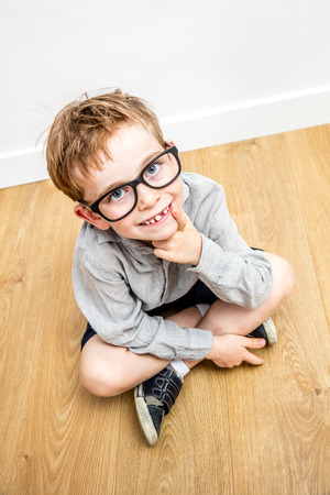 smiling adorable boy with smart eyeglasses and tooth missing seated on the floor thinking, looking up for concept of gifted education and reflection, high angle viewの写真素材