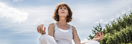breathing outdoors - beautiful 50s woman sitting on a stone in yoga lotus position, seeking for spirituality over summer blue sky,low angle view, long bannerの写真素材