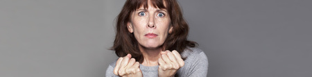 closeup portrait of tensed beautiful 50's woman with both fists held strong for success and stimulation, grey bannerの写真素材