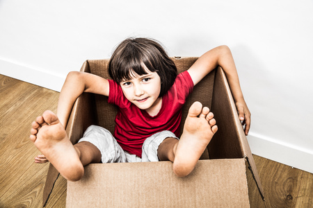 relaxed 6 years old child sitting in an old cardboard box with feet out playing for fun, building toy, moving day or hiding in new house, one real personの写真素材