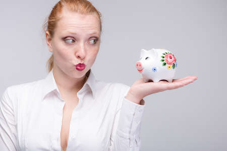 happy young red haired woman wants to kiss her piggy bank / porcelain bank in her handの写真素材