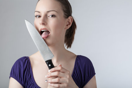 A young woman is licking at a sharp kitchen knifeの写真素材