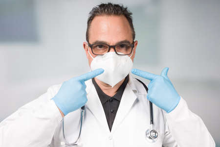 Doctor with medical face mask and medical gloves points to his medical mask in front of a clinic roomの写真素材