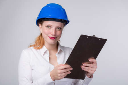 Young female red haired engineer wearing a blue hard hat is working with a clip boardの写真素材