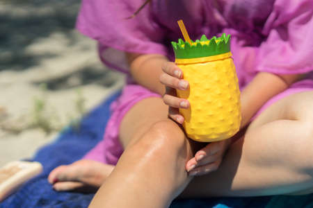 Young woman holding a colorful drink cup on a beach. Sitting in the sand with a pineapple shaped plastic glass, yellow and green color. Girl holding a refreshing beverage in her hands on a sunny day.の写真素材