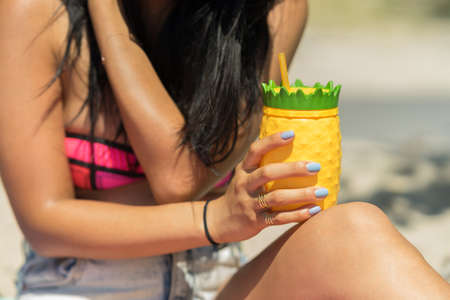 Young woman holding a colorful drink cup on a beach. Sitting in the sand with a pineapple shaped plastic glass, yellow and green color. Girl holding a refreshing beverage in her hands on a sunny day.の写真素材