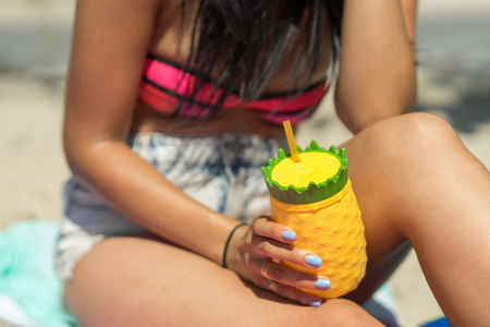 Young woman holding a colorful drink cup on a beach. Sitting in the sand with a pineapple shaped plastic glass, yellow and green color. Girl holding a refreshing beverage in her hands on a sunny day.の写真素材