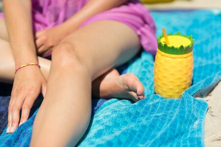 Young woman holding a colorful drink cup on a beach. Sitting in the sand with a pineapple shaped plastic glass, yellow and green color. Girl holding a refreshing beverage in her hands on a sunny day.の写真素材