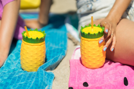 Young woman holding a colorful drink cup on a beach. Sitting in the sand with a pineapple shaped plastic glass, yellow and green color. Girl holding a refreshing beverage in her hands on a sunny day.の写真素材