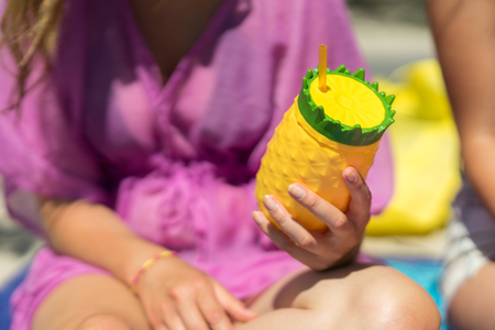 Young woman holding a colorful drink cup on a beach. Sitting in the sand with a pineapple shaped plastic glass, yellow and green color. Girl holding a refreshing beverage in her hands on a sunny day.の写真素材