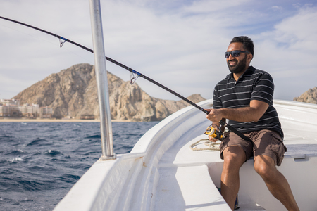 Young East Asian man fishing on open water from the boat. Beautiful sunny day with the shoreline visible in the background.の写真素材