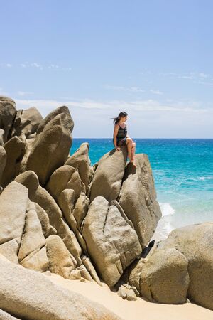 Young woman lounging on the rock formations around the Arch in Cabo San Lucas, Mexico. Interesting shapes of cliffs and stones, beautiful blue water and sunlight. Peaceful vacation spot.の写真素材