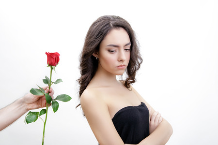 Beautiful upset girl receives one red rose. She looks at the flowers. She is looking over her shoulder. Men's hand holding one rose. Girl is white with bushy brown hair. Isolated on white background.の写真素材