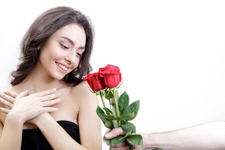 Beautiful girl receives three red roses. She is surprised, looking at the flowers and smiling. Men's hand holding three roses. Girl is white with bushy brown hair. Isolated on white background.の写真素材