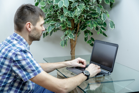 Serious man working at a laptop at home. A man sitting at a glass table. He is dressed in a shirt and jeans. Wristwatch on the hand.の写真素材