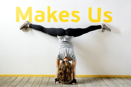 Fit woman doing a handstand near the wall below the phrase "Makes Us". Fitness workout. Concept balance sport fitness. Female in sportswear with long blond hair. View from the back.の写真素材