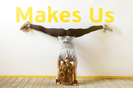 Fit woman doing a handstand near the wall below the phrase "Makes Us". Fitness workout. Concept balance sport fitness. Female in sportswear with long blond hair. View from the back. Sunny effect.の写真素材