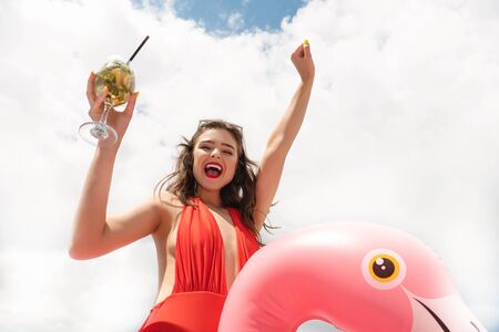 cute sexy young brunette woman with perfect body wearing red swimsuit holding cocktail on pink inflatable flamingo against blue skyの写真素材