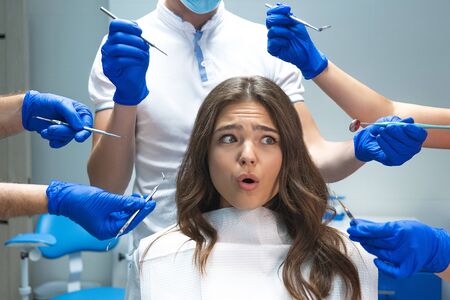 scared young brunette woman patient sitting in dentist chair surrounded by hands in blue gloves with medical instrumentsの写真素材