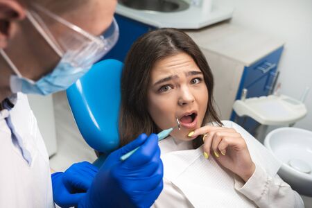 beautiful young scared patient woman having examination at dental office by handsome dentist in mask and blue gloves looks frightenedの写真素材