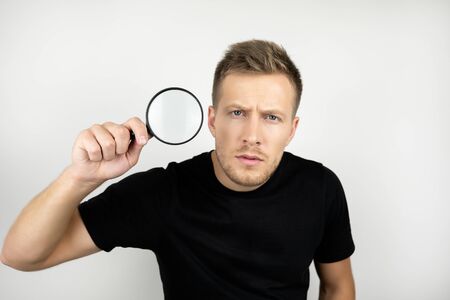 handsome young man wearing black t-shirt holding magnifying glass near his eye looks suspicious...