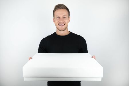 handsome young man wearing black t-shirt holding parcel in paper box for delivery on isolated white...