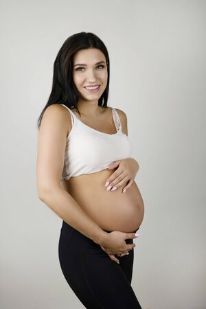 beautiful pregnant brunette woman holding her pregnant naked belly looking content on isolated white...