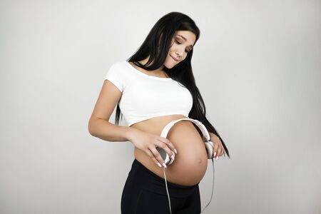 brunette young beautiful pregant woman holding headphones near her belly feeling happy on isolated white backgroundの写真素材