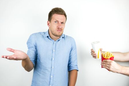 young handsome man hesitates whether to eat fast food french fries and drink soda or not on isolated white backgroundの写真素材