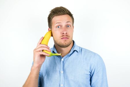 young handsome man holding one fresh banana as a phone near his ear looks suspicious on isolated white backgroundの写真素材