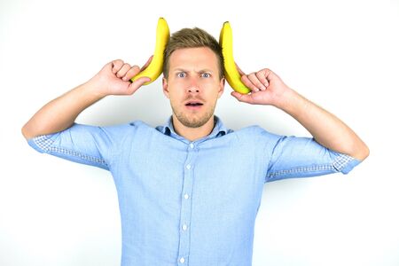 young handsome man holding two fresh bananas near his head looking funny on isolated white backgroundの写真素材