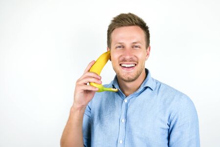 young handsome man holding one fresh banana as a phone near his ear looks happy on isolated white backgroundの写真素材