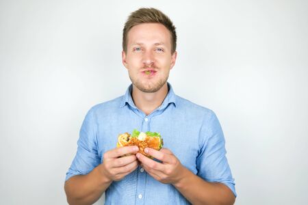 young smiling man biting cheeseburger from fast food restaurant standing with salad leaf in his mouth on isolated white backgroundの写真素材