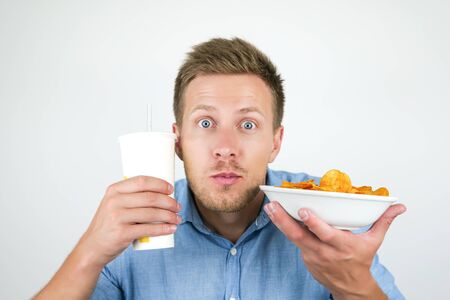 young handsome man looks amazed while holding soda drink and plate with paprika chips near his face on isolated white backgroundの写真素材