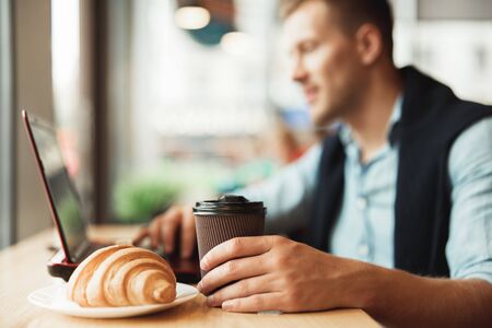 young man working remotely in his laptop eats croissant and holds hot coffee in cafeの写真素材