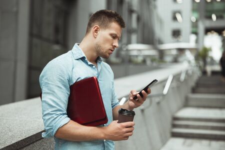 young handsome office worker checking his smarphone holding laptop and cup of coffee standing in the...