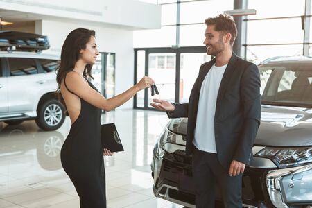 beautiful brunette woman manager giving car keys to handsome man client standing near brand new car...