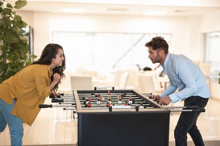two colleagues brunette beautiful woman and handsome young man playing table football during break...