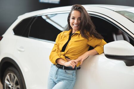 beautiful brunette woman looking for new car in dealership center leaning on white vehicle looking happyの写真素材