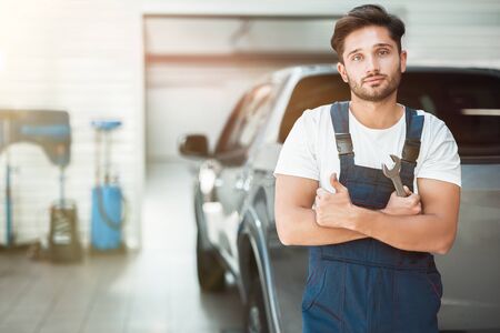 young handsome mechanic wearing uniform holding spanner stands in car service center showing like signの写真素材
