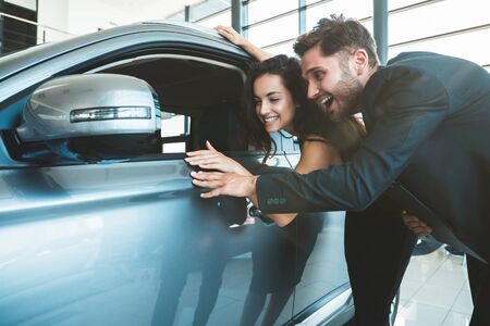 handsome man and young beautiful woman looking happy checking new car interior while choosing new car in dealership centerの写真素材
