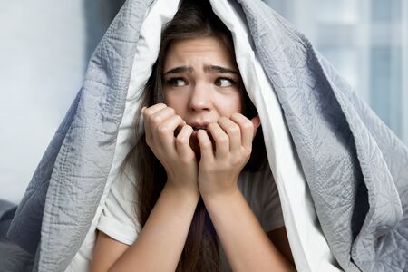 young beautiful brunette woman sitting in bed with blanket on her head looking frightened in the morningの写真素材