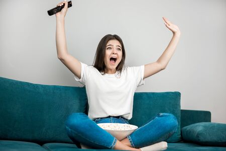 beautiful young woman cheers on her favorite team while watching football game eating popcorn on the sofa at homeの写真素材