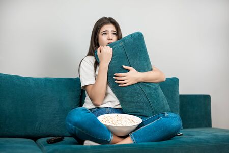 beautiful young woman watching scary movie on the sofa eating popcorn on the sofa at home looking frightened holding pillow in both handsの写真素材