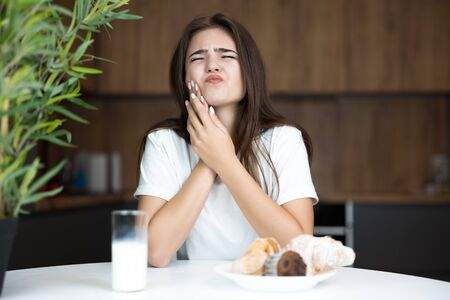 beautiful young woman suffering terrible toothache while eating cupcakes and croissants and drinking milk for breakfast in the kitchen looking sickの写真素材