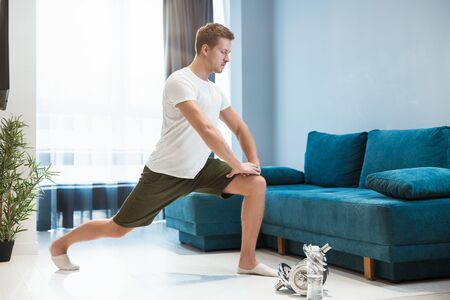 young handsome man doing lunges during stretching before workout at home looking focused sporty and healthy lifestyleの写真素材