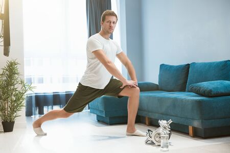young handsome man doing lunges during stretching before workout at home looking happy sporty and healthy lifestyleの写真素材